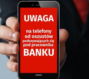 Telefon komórkowy w dłoni, a na ekranie telefonu napis &quot;Uwaga na telefony od oszustów podszywających się za pracownika banku&quot;.